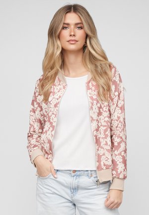 Młoda kobieta o długich blond włosach, ubrana w różową kurtkę bomber z kwiatowym wzorem, białą koszulę oraz jasnoniebieskie jeansy, z jedną ręką w kieszeni.