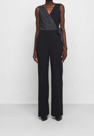 Personne portant une combinaison noire sans manches avec un détail drapé à motif quadrillé et un lien sur un côté, associée à des chaussures pointues noires.