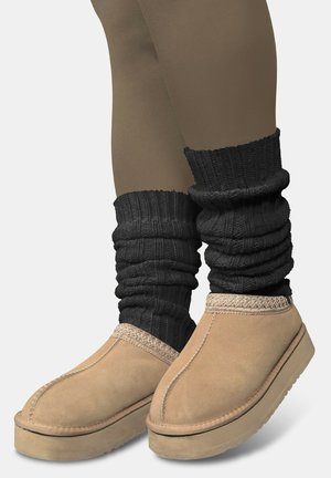 Wildleder-Hausschuhe in Hellbraun mit einer strukturierten Sohle, ergänzt durch schwarze gestrickte Beinstulpen mit gerafftem Muster.