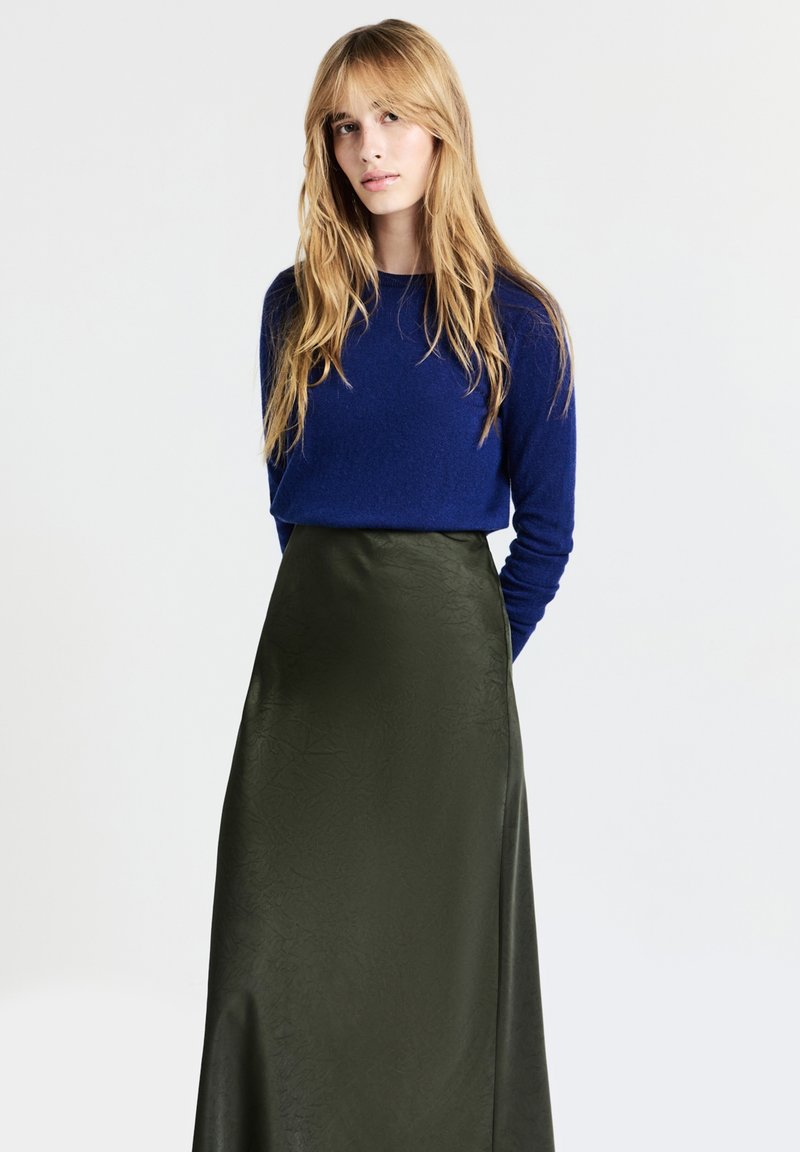 Blonde Frau, die ein navyblaues Langarmoberteil trägt, das in einen hoch taillierten satinmäßigen Rock in dunklem Olivgrün gesteckt ist, steht vor einem schlichten weißen Hintergrund.