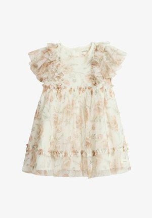 Lille pige med mørkt hår iført en hvid blomsterprintet kjole med flæser og en hvid sommerfugle-hårclips, som smiler let.