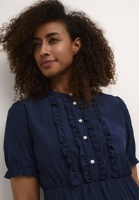 Abito blu navy con maniche a sbuffo corte, design frontale arricciato e bottoni decorativi in perla lungo il centro. Tessuto morbido.
