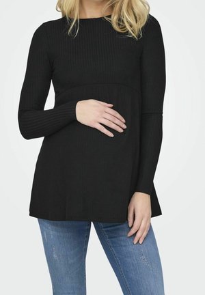 Femme portant un haut noir à manches longues et un jean bleu, tenant doucement sa main sur son ventre.