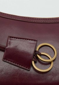 Detaliu al unei genți din piele burgundă, cu o textură netedă, accesorii circulare de culoare aurie și un tab cusut cu o mică pată rectangulară.