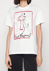 Bílý bavlněný tričko s módním skicováním, červenými akcenty, černým ručně psaným textem a různými siluetami šatů. Pohodlný střih.