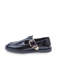 Mocasines de charol negro con detalle de hebilla en tono dorado, punta redondeada y costuras en contraste a lo largo de la suela.
