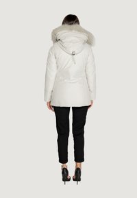 Parka bianca isolata con cappuccio foderato in pelliccia, doppia zip e cinturini regolabili. Abbinata a pantaloni neri slim-fit e tacchi.