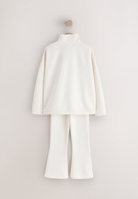 Haut blanc à col roulé manches longues avec poignets élastiques et pantalon large assorti suspendu à un crochet en bois contre un mur beige.