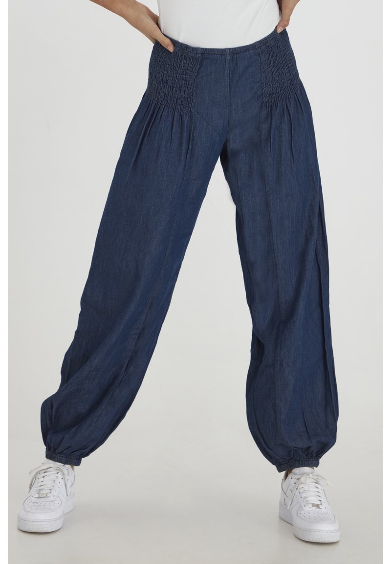 Personne portant un pantalon ample en denim bleu à taille et chevilles élastiquées, baskets blanches et haut blanc sur un fond uni.