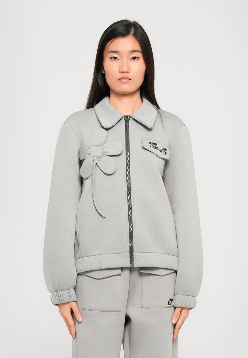 Grå zip-up jakke med stor krave, dekorativ blomsterdetalje og ribbede manchetter. Designet har et logo-patch og en glat tekstur.