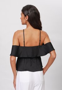 Svart off-shoulder topp med rynket halslinje, tynne justerbare stropper og teksturert stoff. Brukt med hvite bukser, som viser en uformell stil.