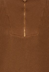 Tissu tricoté brun avec une finition texturée, présentant un design à demi-zip et une petite poche poitrine bordée de côtes. Détail de fermeture éclair dorée.