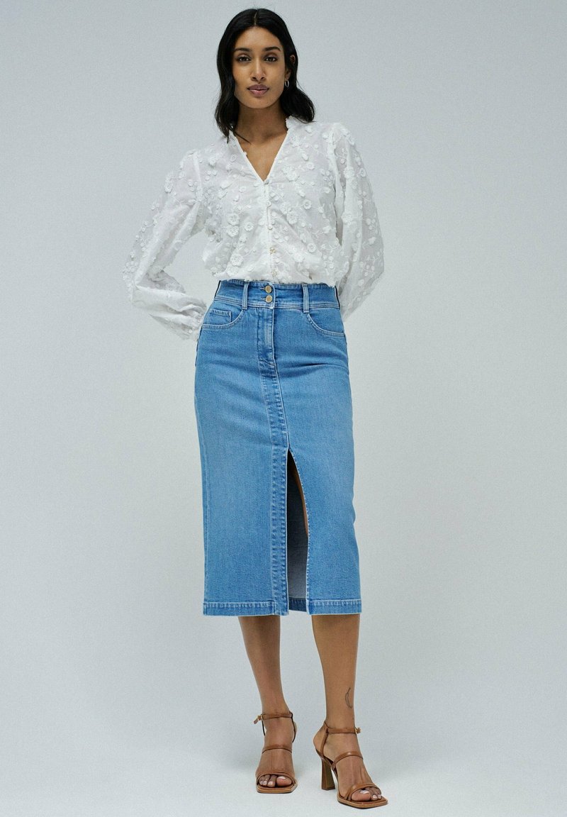 Blusa ligera blanca de manga larga con diseño floral texturizado. Falda midi de mezclilla azul con abertura frontal y dos botones en la cintura. Sandalias de tacón marrones.