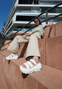 Weiße Plattformsandalen mit drei breiten Riemen, ausgestattet mit einem geschwungenen Sohlen-Design und einer strukturierten Fußbett, vor einem rötlichen Betonhintergrund.