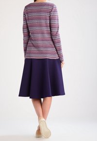 Dzianinowy pulower z różowo-fioletowym wzorem w zygzaki, zestawiony z ciemnofioletową, rozkloszowaną spódnicą. Widok od tyłu, pokazujący casualowe obuwie.