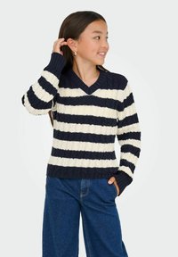 Mergina su ilgais tamsiais plaukais, dėvinti auksinius auskarus-žiedus, tamsiai mėlyną ir baltą juostuotą megztinį bei mėlynas kelnes, pozuoja su viena ranka arti ausies.