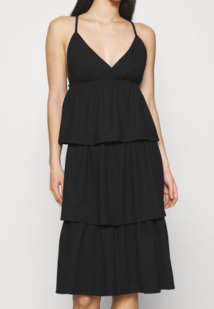 Musta kerroksittainen mekko V-pääntiellä, ohuilla olkaimilla ja teksturoidulla kankaalla. Mekossa on kolme kerrosta, jotka luovat virtaavan siluetin.