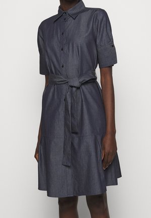 Robe-chemise manches courtes en denim foncé avec col, boutonnée sur le devant et ceinture à la taille, portée par une personne à la peau foncée sur un fond uni.