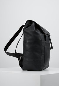 Svart läderryggsäck med strukturerad yta, rulltoppsdesign, justerbar rem och sidofickor. Har metalliska detaljer som accenter.