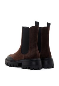 Botas de tobillo de ante marrón que presentan paneles laterales elásticos, una puntera negra contrastante y una suela de goma negra gruesa con relieves.