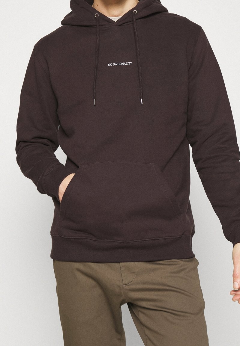 Ciemnobrązowa bluza z kapturem, przednią kieszenią kangurką, regulowanym sznurkiem i białym napisem "NO NATIONALITY" na piersi.