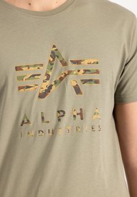 Alyvuogių spalvos medvilninis marškinėliai su dideliu kamufliažo rašto logotipu ir kontrastinga užrašu „ALPHA INDUSTRIES“. Apvalus kaklas.