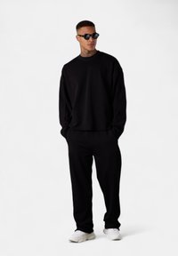 Mann in schwarzem oversized Sweatshirt und Hose, weißen Sneakers, schwarzen Sonnenbrille, mit Tätowierungen am Hals und den Ohren, steht vor einem weißen Hintergrund.