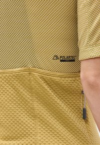 Gros plan sur un tissu jaune texturé d'une veste avec le logo Polartec Delta et une poche zippée près de la couture latérale.