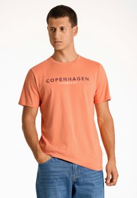 Kortärmad orange t-shirt med texten "COPENHAGEN LINDENBERGH" tryckt i mörklila. Tillverkad av slät tyg, kombinerad med blå denimjeans.