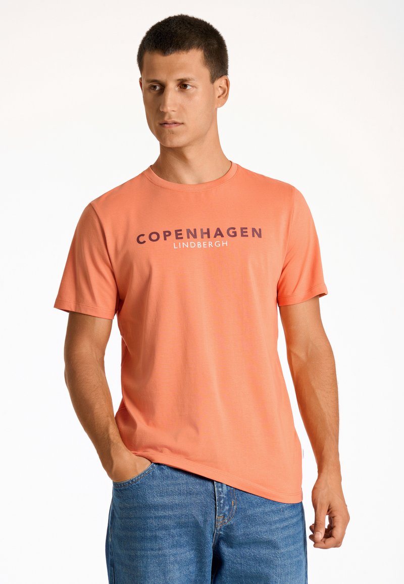 Kortärmad orange t-shirt med texten "COPENHAGEN LINDENBERGH" tryckt i mörklila. Tillverkad av slät tyg, kombinerad med blå denimjeans.
