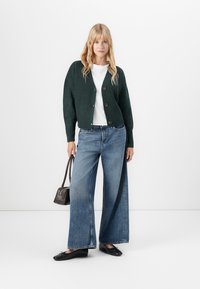 Kvinne står iført en mørkegrønn cardigan, hvit topp, vide blå jeans, svarte leiligheter, og holder en liten mørk håndveske.