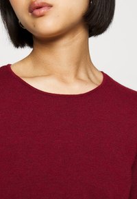 Maglione in maglia bordeaux con collo rotondo. La texture è liscia e finemente lavorata, con punti visibili lungo il bordo del colletto.