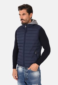 Gilet in stile puffer blu navy con chiusura a zip, design trapuntato e cappuccio grigio. Caratterizzato da tasche laterali e una silhouette aderente. Indossato sopra una maglietta a manica lunga nera.