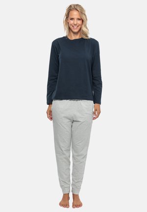 Kvinna som står barfota iförd en marinblå långärmad tröja och ljusgrå sweatpants, leende mot en enhetlig ljus bakgrund.