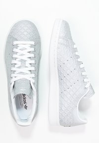 Zapatillas deportivas grises con un patrón texturizado de escamas, cordones blancos y una suela de goma blanca. El interior muestra una etiqueta de la marca.