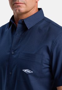 Dunkelblaue Hemd mit Knöpfen, das einen spitzen Kragen, eine linke Brusttasche mit gesticktem Logo und eine glatte Baumwollstoffstruktur aufweist.