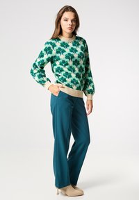 Grüner, blattmusterbestickter Strickpullover mit langen Ärmeln und cremefarbigem Rippenkragen. Türkisfarbene, weite Hosen kombiniert mit beigen, hohen Stiefeletten.