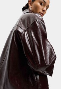 Burgundy imitatkläde jacka med blank yta, oversize passform och strukturerade axlar. Har en spetsig krage och sömnadsdetaljer.