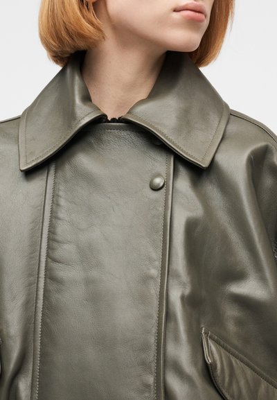 Γυναίκα με κοντά κόκκινα μαλλιά που φο ράει γκρι-πράσινο δερμάτινο μπουφάν με φαρδιά γιακά και κουμπιά με τρουκ, εμφανίζεται από τον λαιμό μέχρι το στήθος.