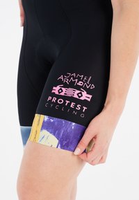 Zwarte fietsshorts met een gevoerde inzet, met roze logo's "JAMEL ARMAND" en "PROTEST CYCLING." Kleurrijke, gemusterde zoom met paars en geel.