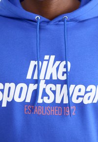Blå huvtröja med fet vit text som läser "nike sportswear" med "ETABLERAD 1972" i orange. Inkluderar snoddar och mjuk textur.