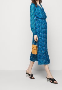 Vestido azul con estampado, de manga larga y con dobladillo con volantes, combinado con sandalias negras de tacón y un pequeño bolso amarillo. Tejido suave y ligero.