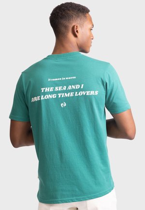Teal cotton t-shirt featuring a round neck, short sleeves, and printed text on the back: "it comes in waves" and "THE SEA AND I ARE LONG TIME LOVERS."