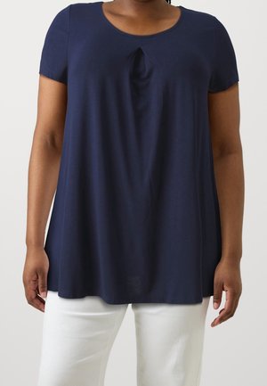 Kobieta ubrana w granatową tunikę z krótkim rękawem i plisą z przodu oraz białe spodnie, stojąca na jednolitym tle.