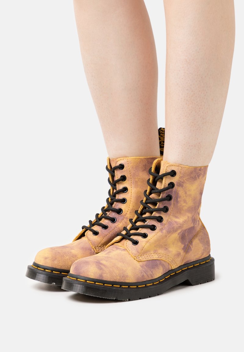 Paire de bottes à lacets jaune moutarde usées avec semelles et coutures noires, portées sur des jambes nues devant un fond blanc.
