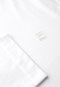 Camiseta de algodón blanca con un pequeño logotipo en relieve en el área del bolsillo del pecho, con una textura suave y lisa y bordes limpios.