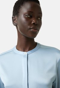 Blouse bleu clair à col, en tissu lisse, avec une fermeture à boutons et de subtiles détails de couture le long des épaules.