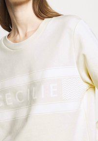 Ljusgul sweatshirt gjord av mjukt tyg med rund halsringning. Har en grafisk tryck med namnet "CECILIE" och ett zickzack-mönster.