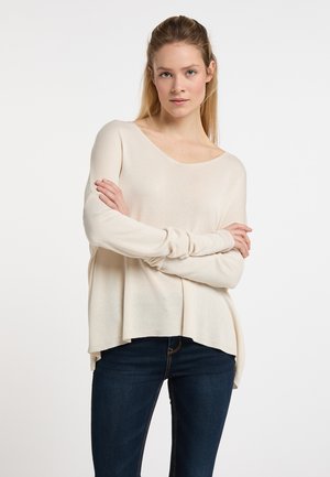 Crème kleurige, losse gebreide trui met een ronde halslijn en lange mouwen, voorzien van een gebogen zoom. Gecombineerd met donkerdenim jeans.