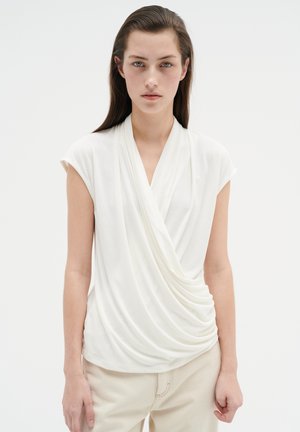 Top blanco con mangas cortas tipo capucha, tejido de jersey suave y un diseño fruncido en el costado, combinado con pantalones beige.